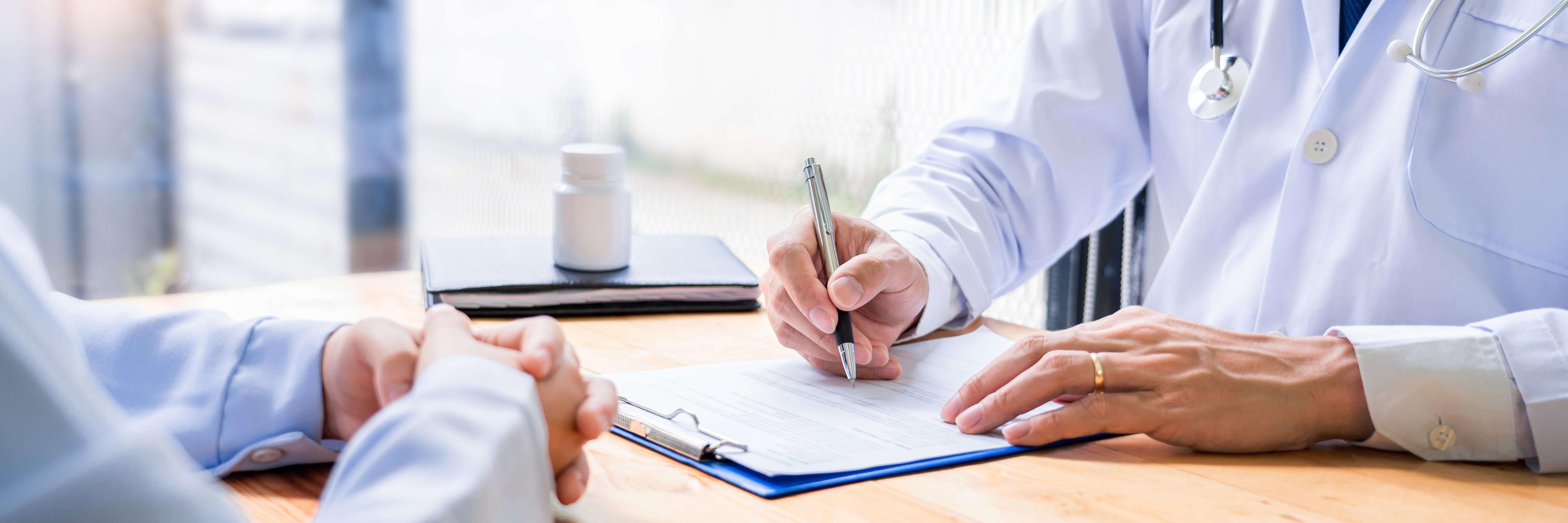 Profissional de saúde com jaleco escrevendo em um formulário, enquanto outro paciente aguarda à frente na mesa.